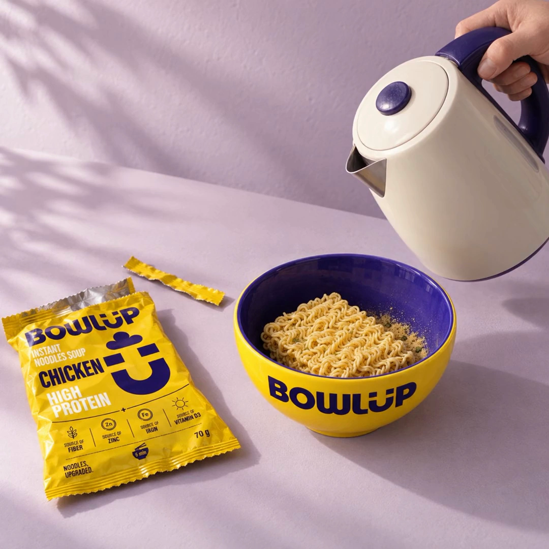 An empty, torn BOWLUP Chicken wrapper lies beside a yellow BOWLUP bowl of dry rectangular noodles, with a kettle ready to pour.